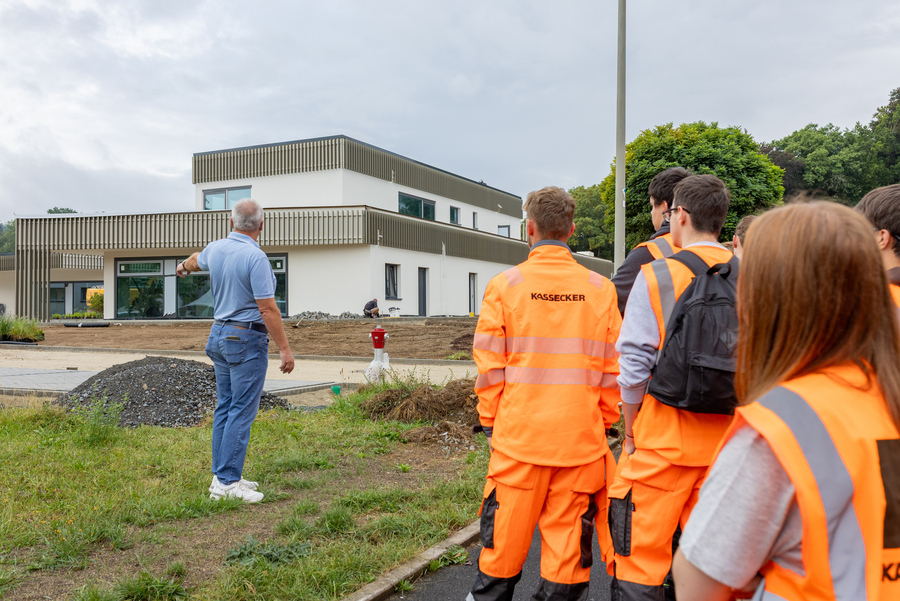Kassecker-Auszubildende stehen in oranger Warnkleidung vor einem zweistöckigen, modernen Neubau. Ein Mann in Jeans und blauem Shirt deutet auf die noch nicht fertiggestellten Außenanlagen