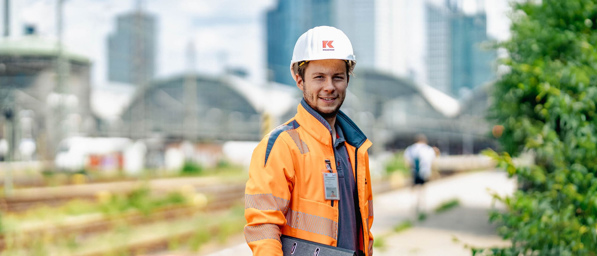Bauleiter der Unternehmensgruppe Kassecker an der Eisenbahnstrecke Bauarbeiter in auffälliger Kleidung hält ein Klemmbrett neben Eisenbahnschienen, mit Industrieanlagen und Wolkenkratzern im Hintergrund.