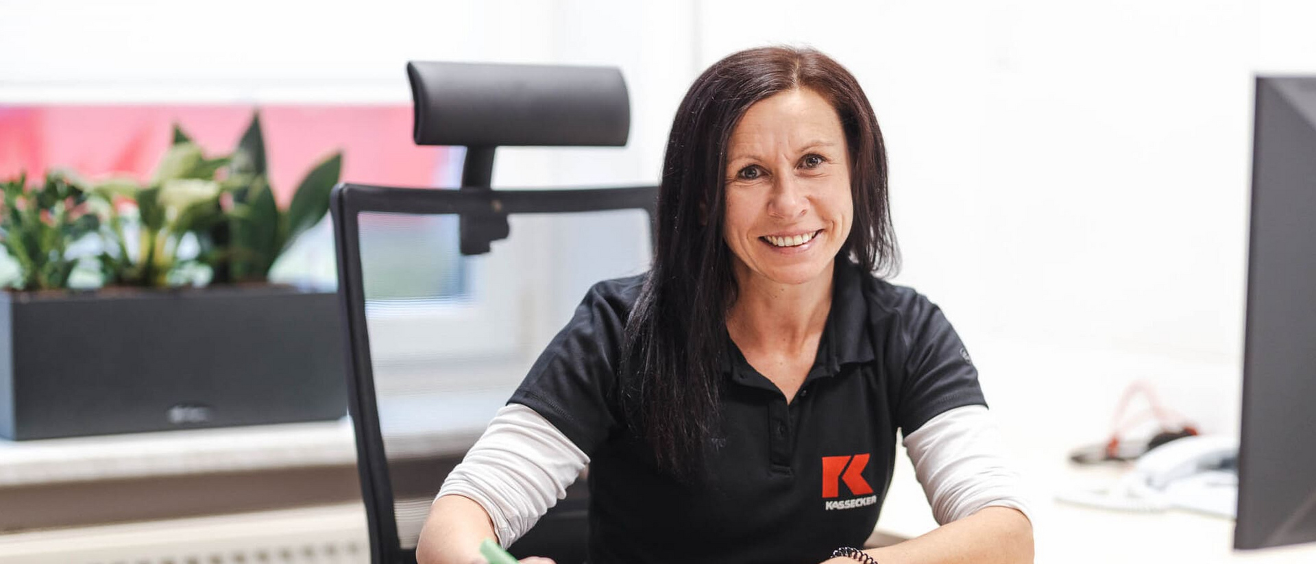 Büroangestellte bei KASSECKER Eine lächelnde Frau sitzt an einem Schreibtisch in einem Büro und hält einen Stift in der Hand. Auf ihrem schwarzen Hemd ist das Logo der Firma KASSECKER zu sehen.