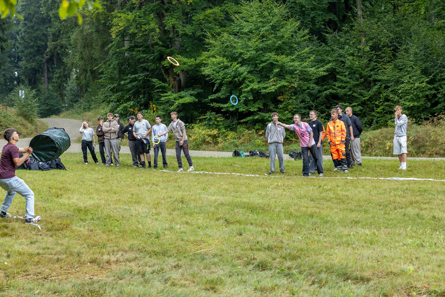 Eine Grupe Jugendlicher steht auf einer Wiese. Einige Meter weiter auf der Wiese steht ein einzelner Jugendlicher. Er versucht einen von der Gruppe aus geworfenen Ring mit einem Gartenbeutel aufzufangen.